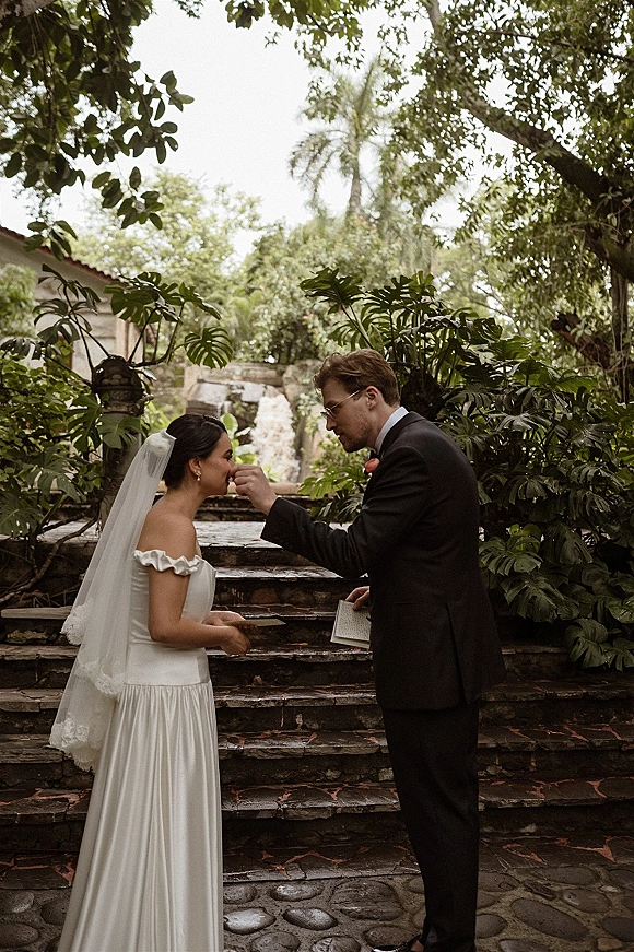 Wedding vows as groom reads from a vow book to veiled bride in off-the-shoulder dress on stone steps amid tropical greenery courtyard