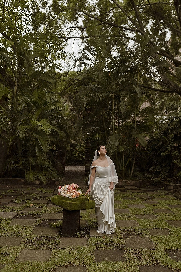 Bridal portrait of a bride in an off-the-shoulder satin wedding dress with veil, holding a peach rose bouquet in a tropical palm garden