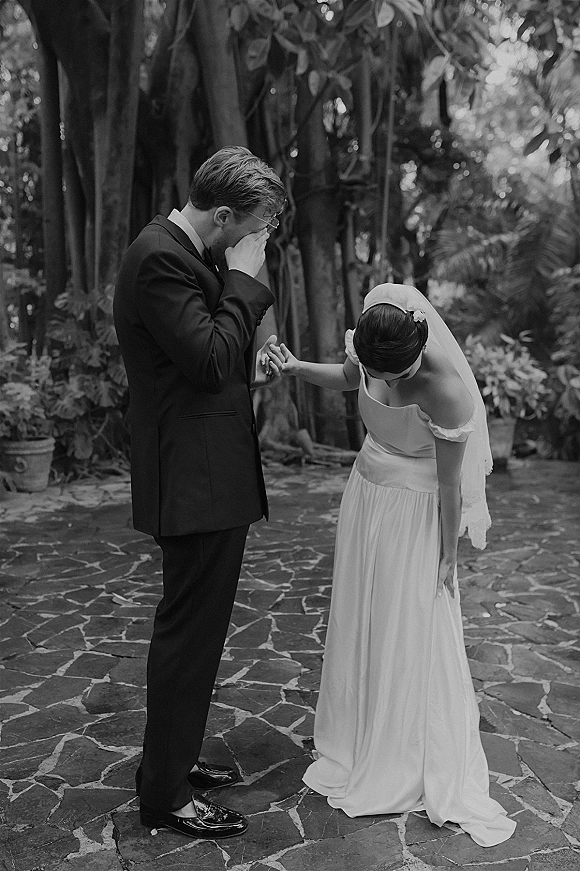 Wedding first look as the groom cries, wiping tears while holding the bride’s hand on a stone patio among garden trees and potted plants