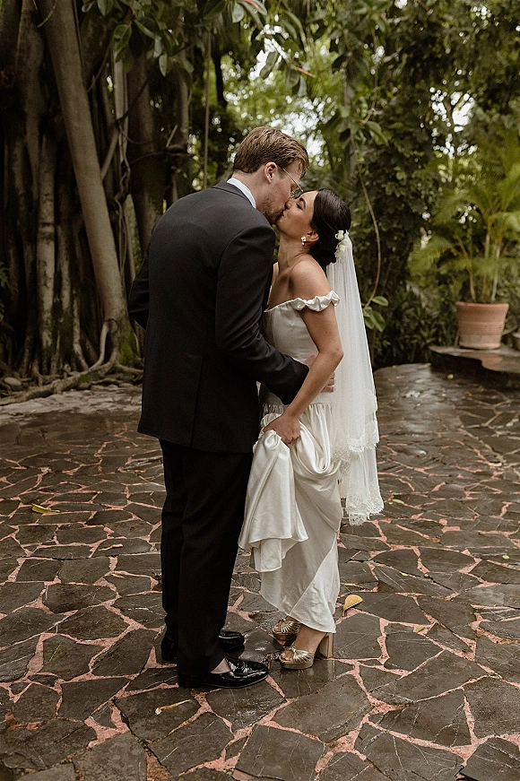 Wedding kiss as the bride and groom kissing on a stone patio, her veil and off-shoulder dress framed by tropical greenery and roots