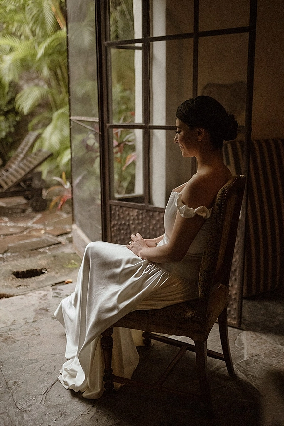 Bridal portrait of a bride sitting by window in a strapless satin wedding dress, bridal updo and ring visible, by open panes with tropical greenery outside
