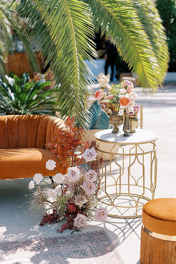 Wedding lounge seating with a velvet sofa, gold round side table, and lush florals on a sunlit patio with trees in back