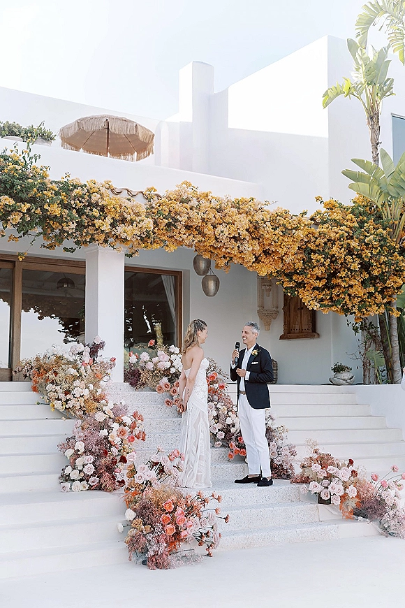 Wedding vows as the groom speaks into a microphone beside the bride on flower-lined villa steps with hanging lanterns on the terrace