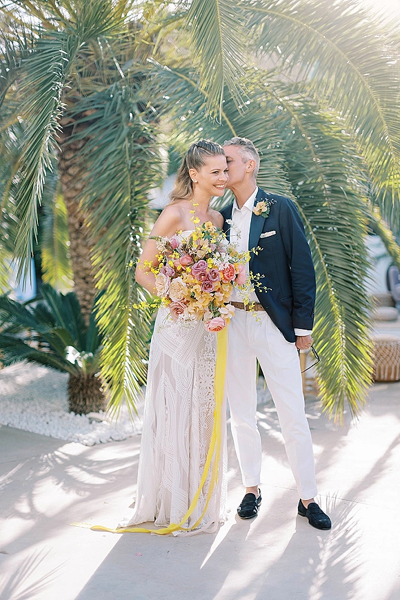 Couple portrait with groom kissing bride’s forehead as she holds a colorful bouquet, sun flare glowing behind palm trees on a patio