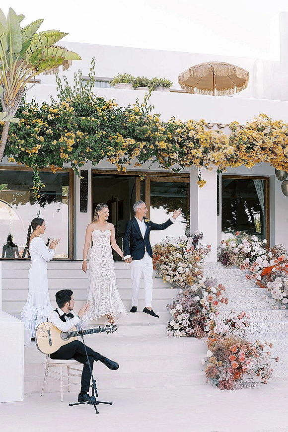 Ceremony moment as bride and groom entrance holding hands on villa steps, with blush florals, hanging flowers and microphone setup