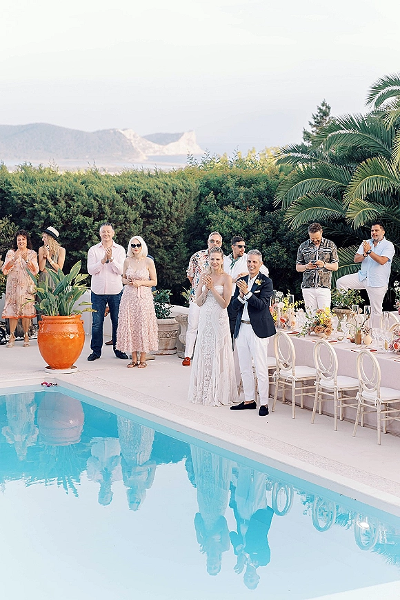 Wedding reception moment as bride in strapless lace dress and groom in navy blazer receive applause beside a poolside table with taper candles