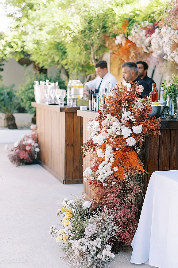 Wedding bar setup with a wooden outdoor wedding bar, lush florals and greenery, glassware, liquor bottles, shakers, and lemons on a garden patio