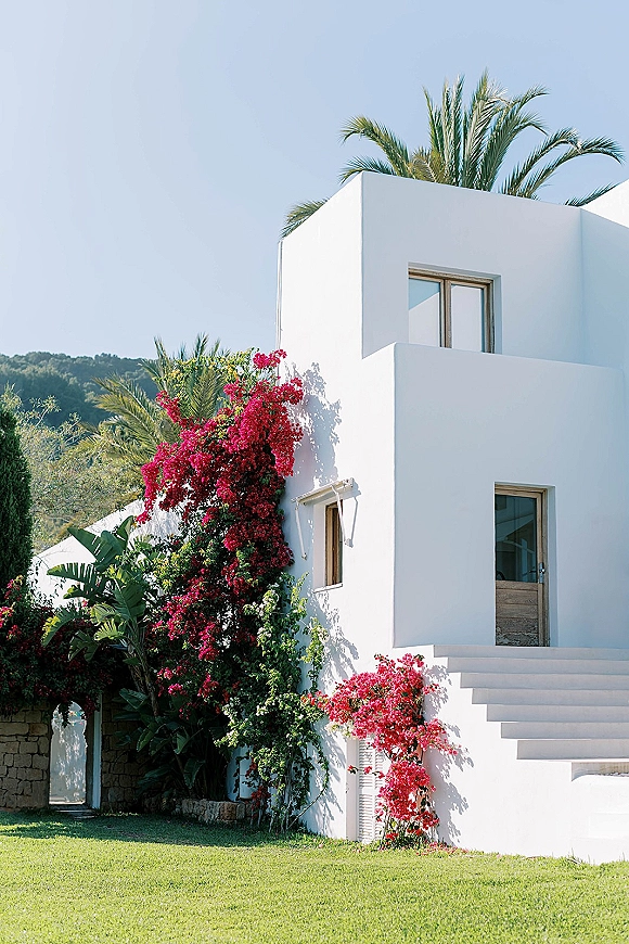 White wedding venue with bougainvillea vines on a modern villa facade, framed by palm trees, stone wall, and stairs under blue sky