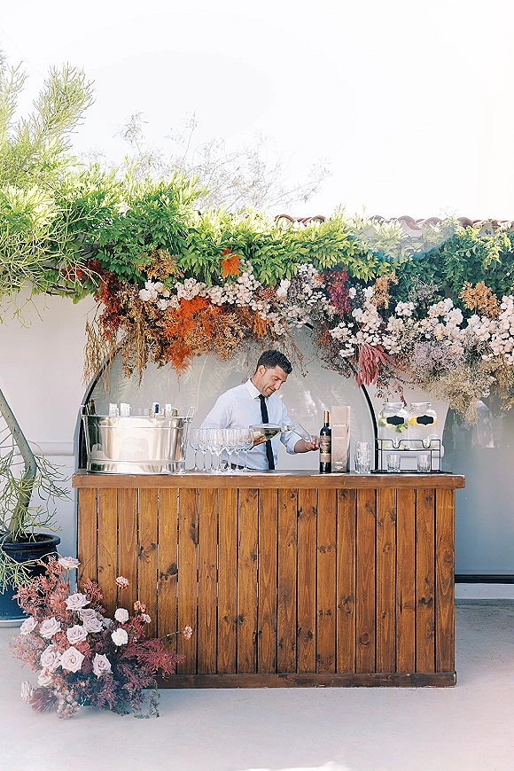 Wedding bar setup with a rustic wedding bar, wood counter and floral installation, champagne flutes and wine bottles on a patio by a white wall