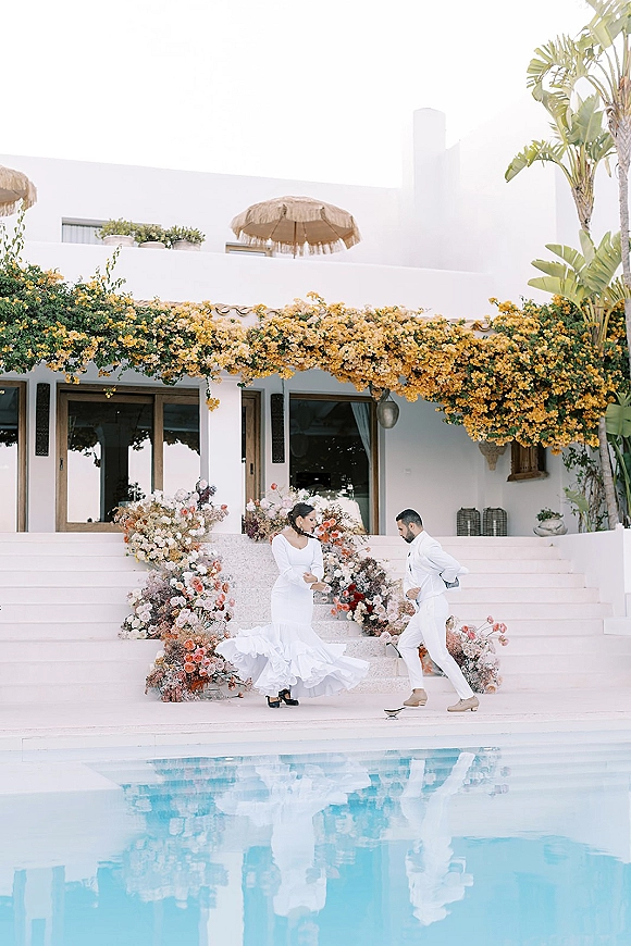 Couple portrait of bride twirling her long-sleeve gown beside groom in white suit by a villa pool, with floral staircase decor