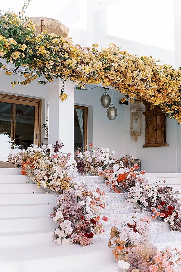 Wedding entrance florals and wedding staircase flowers cascade over porch steps with pastel roses, dried blooms, lanterns, and a thatched umbrella backdrop