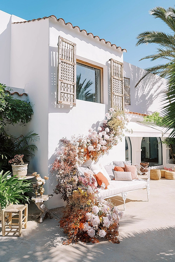 Lounge seating decor with an outdoor sofa, throw pillows, woven poufs, and candle holders beneath a floral installation on a stucco patio