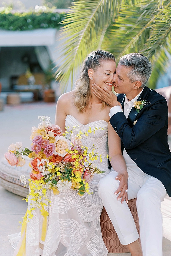 Wedding couple portrait of groom kissing bride’s cheek as he cradles her face, bouquet with roses visible on a palm-lined patio