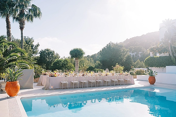 Poolside reception setup with long banquet table, white linens, clear chairs, colorful florals and taper candles beside a palm-lined pool and hills