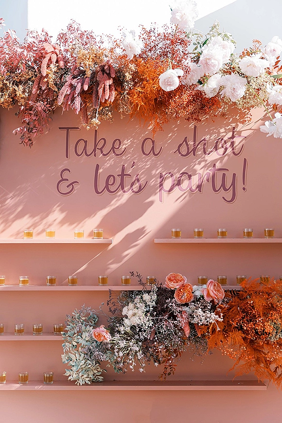 Shot wall display with wedding shot wall shelves of glasses, take a shot sign, and orange-white dried florals against a pink backdrop in sunlight