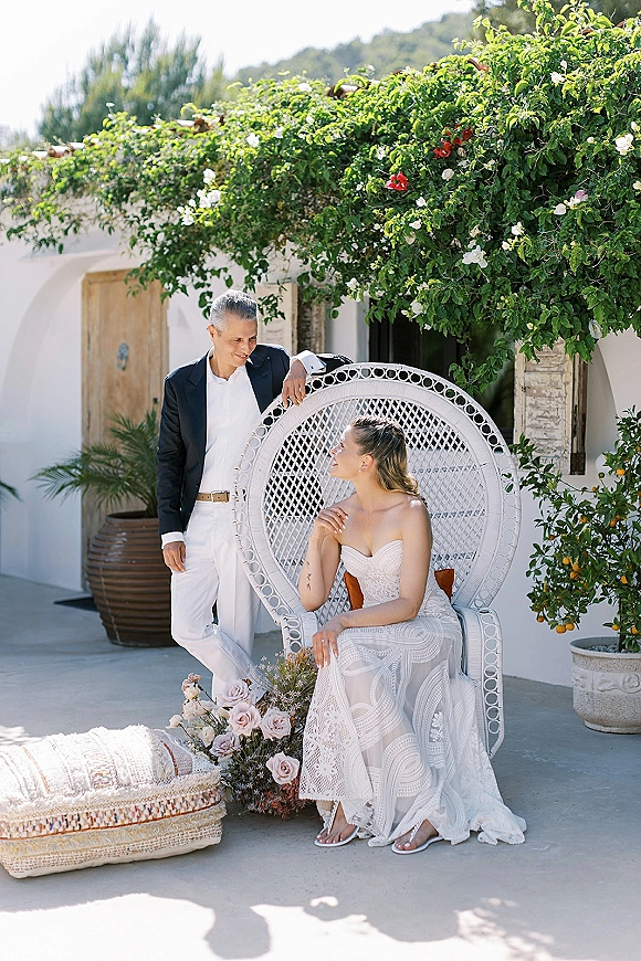 Couple portrait of bride in a strapless lace dress seated in a peacock chair, groom in white pants beside her on a vine-lined patio