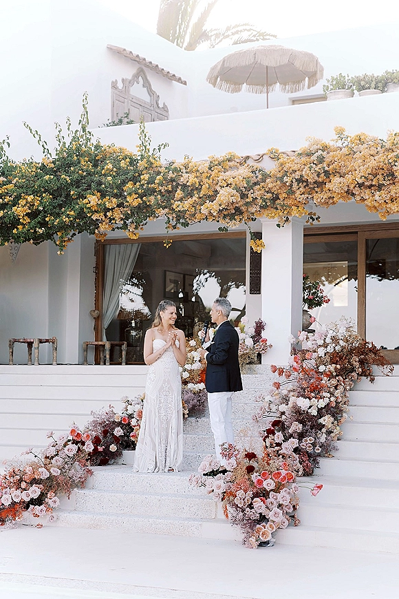 Ceremony vows as groom reads into a microphone beside bride in a strapless lace dress on villa steps with blush florals and pampas grass