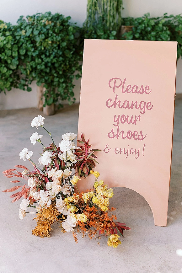 Wedding signage with script lettering on a freestanding sign board, with a peach floral arrangement and greenery against a white wall
