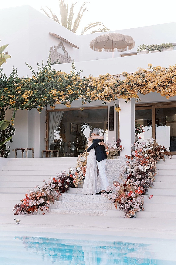 Couple portrait of bride and groom hug on villa steps beside pink and peach wedding florals, with pool and palm tree behind them
