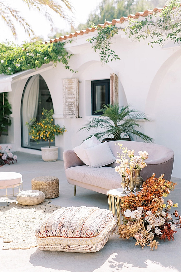 Wedding lounge area with a curved sofa, throw pillows, and gold side table styled with florals under white stucco arches on a patio