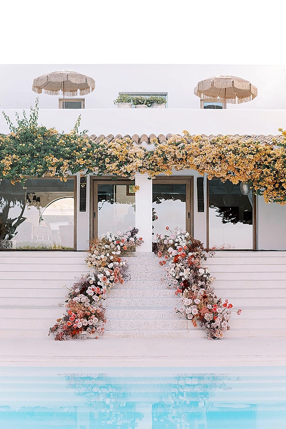 Ceremony backdrop with floral staircase decor of roses and greenery framing villa steps by a pool, with patio umbrellas and balcony above