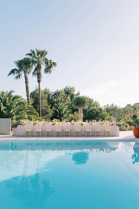 Reception tablescape with long banquet table wedding styling, candles and floral centerpieces beside a pool on a palm-lined patio