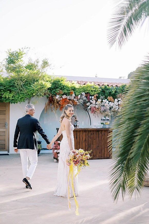 Couple portrait with bride looking back, holding hands as they walk in a palm-lined courtyard, bride carrying bouquet with ribbon streamers