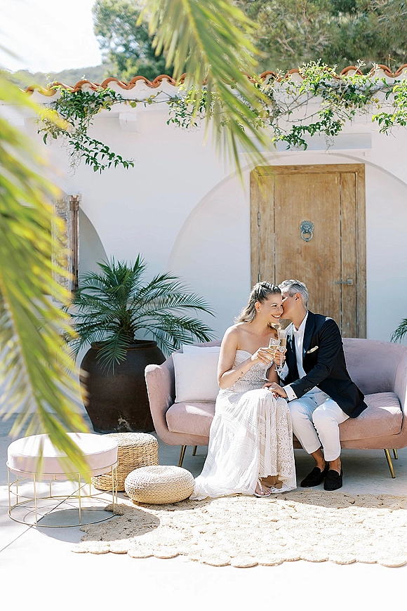 Couple portrait during a wedding champagne toast, bride in strapless lace dress and groom in dark jacket seated on outdoor sofa by arched doorway