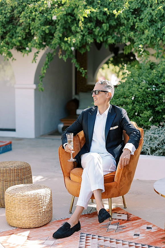 Groom portrait in sunglasses seated in a lounge chair, holding champagne on an outdoor patio with greenery and an arched doorway backdrop