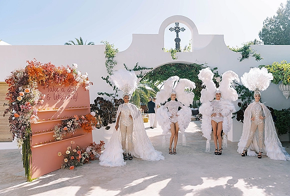 Wedding entertainers in sequined bodysuits with feather headdresses pose by a floral escort wall with candles under a white stucco archway outdoors