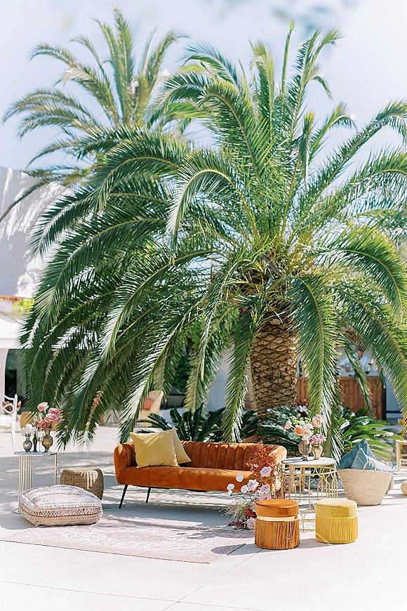 Wedding lounge seating with a velvet sofa, pillows, and poufs around an area rug, styled with florals and lanterns in a palm courtyard patio