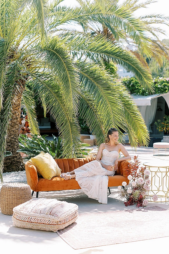 Bridal portrait of a bride lounging on a velvet sofa in a strapless lace wedding dress, with palm trees and patio greenery behind