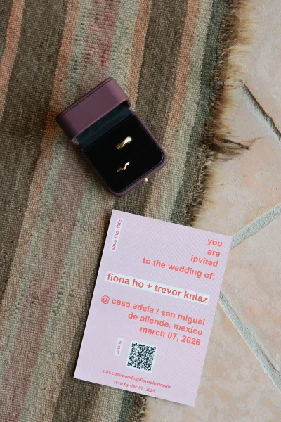 Wedding invitation and save the date card with QR code beside gold wedding bands and ring box on a woven rug over tile floor