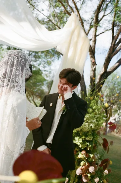 Wedding vows as bride reads from a vow booklet to a teary groom beneath a draped arch, long lace veil and florals outdoors