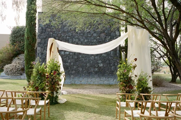Ceremony altar decor with outdoor wedding altar draped fabric and floral pillars, set before a stone wall with wooden chairs on grass