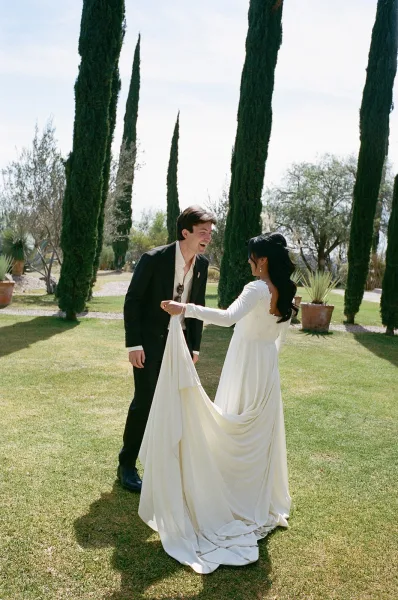 Couple portrait of bride holding her long dress train and groom in a black suit, laughing on a lawn with tall cypress trees behind