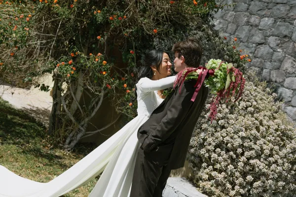 Couple portrait of bride and groom embracing, her long sleeve wedding dress and bouquet with burgundy ribbon against a sunlit stone wall with vines