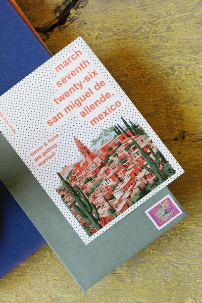Wedding invitation with illustrated cityscape and cactus, dotted card and envelope with postage stamp on blue paper over wood table