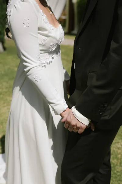 Wedding couple portrait of bride and groom holding hands, lace appliqué long-sleeve gown and black suit on a sunlit lawn with blurred guests