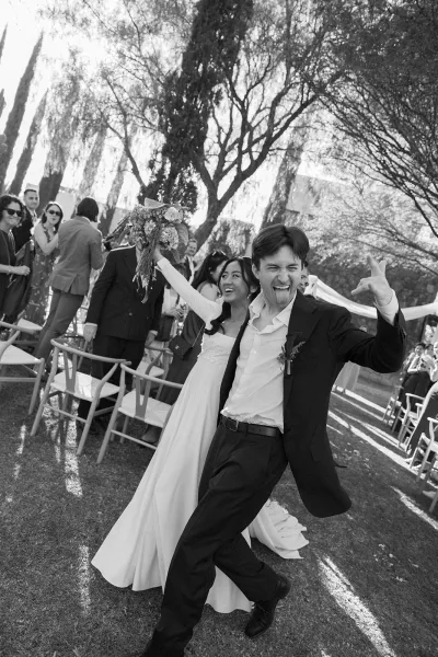 Wedding recessional couple walking down aisle as bride laughs with bouquet raised overhead and groom flashes a rock hand sign on a sunny lawn