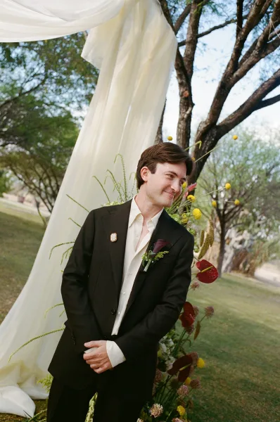 Groom portrait in a black suit with anthurium boutonniere and lapel pin, standing by a draped floral ceremony backdrop outdoors