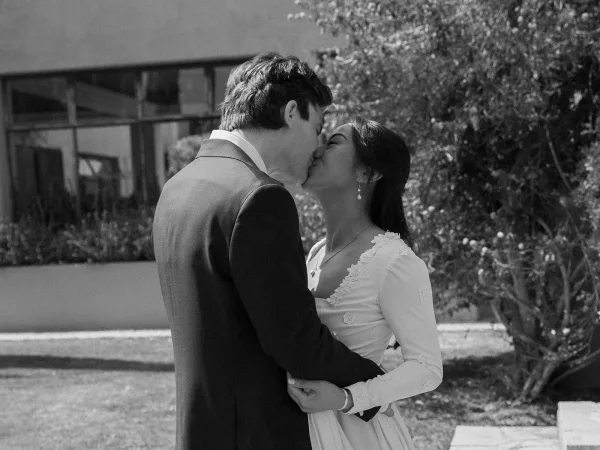 Wedding kiss as bride in long sleeve gown and groom in suit embrace on a garden walkway with trees and building behind