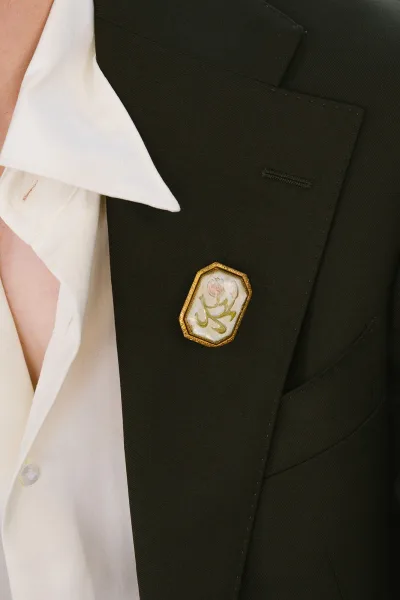 Groom boutonniere with a gold boutonniere pin featuring a floral motif on a black tuxedo lapel and crisp white shirt collar