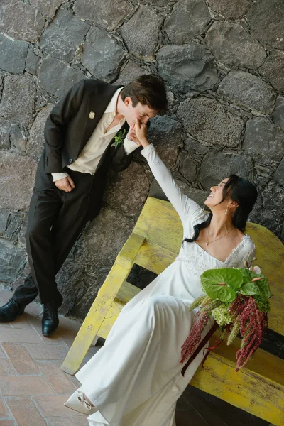Couple portrait of groom kissing bride’s hand as she sits on a yellow bench, holding a cascading anthurium bouquet by a stone wall