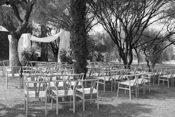 Ceremony setup with wood chairs arranged on a grass lawn facing a fabric-draped arch with floral arrangements in a tree-lined garden