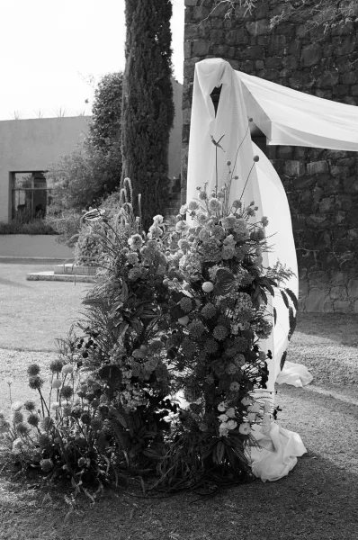 Wedding ceremony arch with a floral wedding arch design, draped white fabric and greenery set against a stone wall in a garden lawn setting