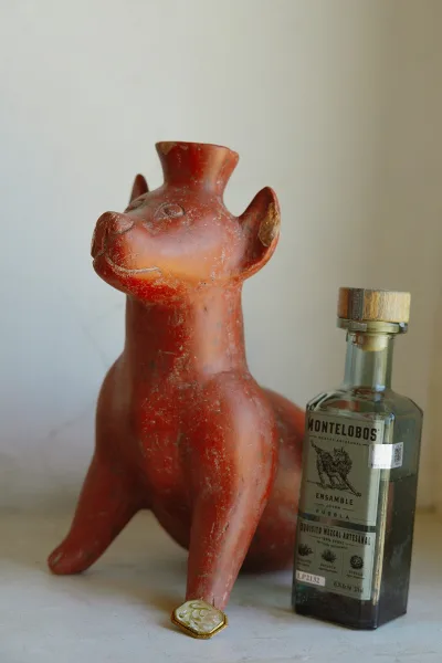 Groom accessories flat lay with a cologne bottle, mezcals bottle, and clay animal figurine arranged on a tabletop against a neutral wall