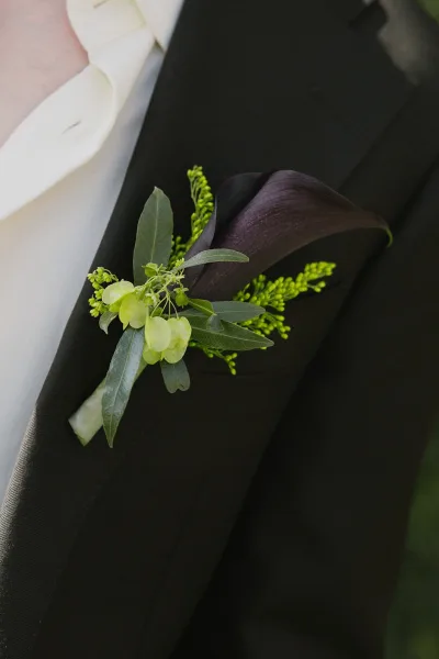 Groom boutonniere with calla lily wrapped in greenery and ribbon on a tuxedo lapel, set against dark suit fabric and soft outdoor blur