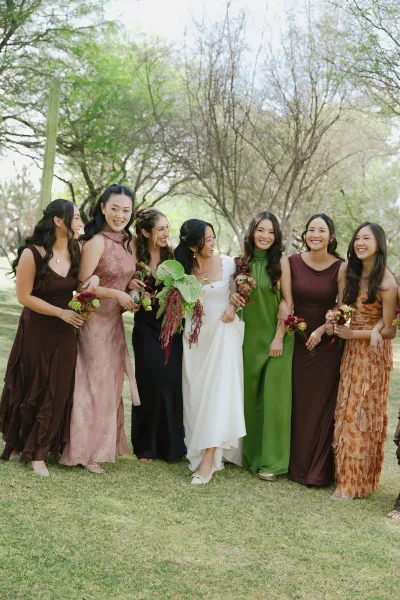 Bridesmaid group photo with bride with bridesmaids laughing arm in arm, holding bouquets on a grassy lawn with trees and cactus backdrop