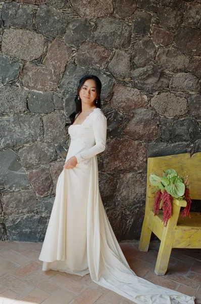 Bridal portrait of a bride in a long sleeve wedding dress with train, looking over her shoulder by a stone wall, bench with florals